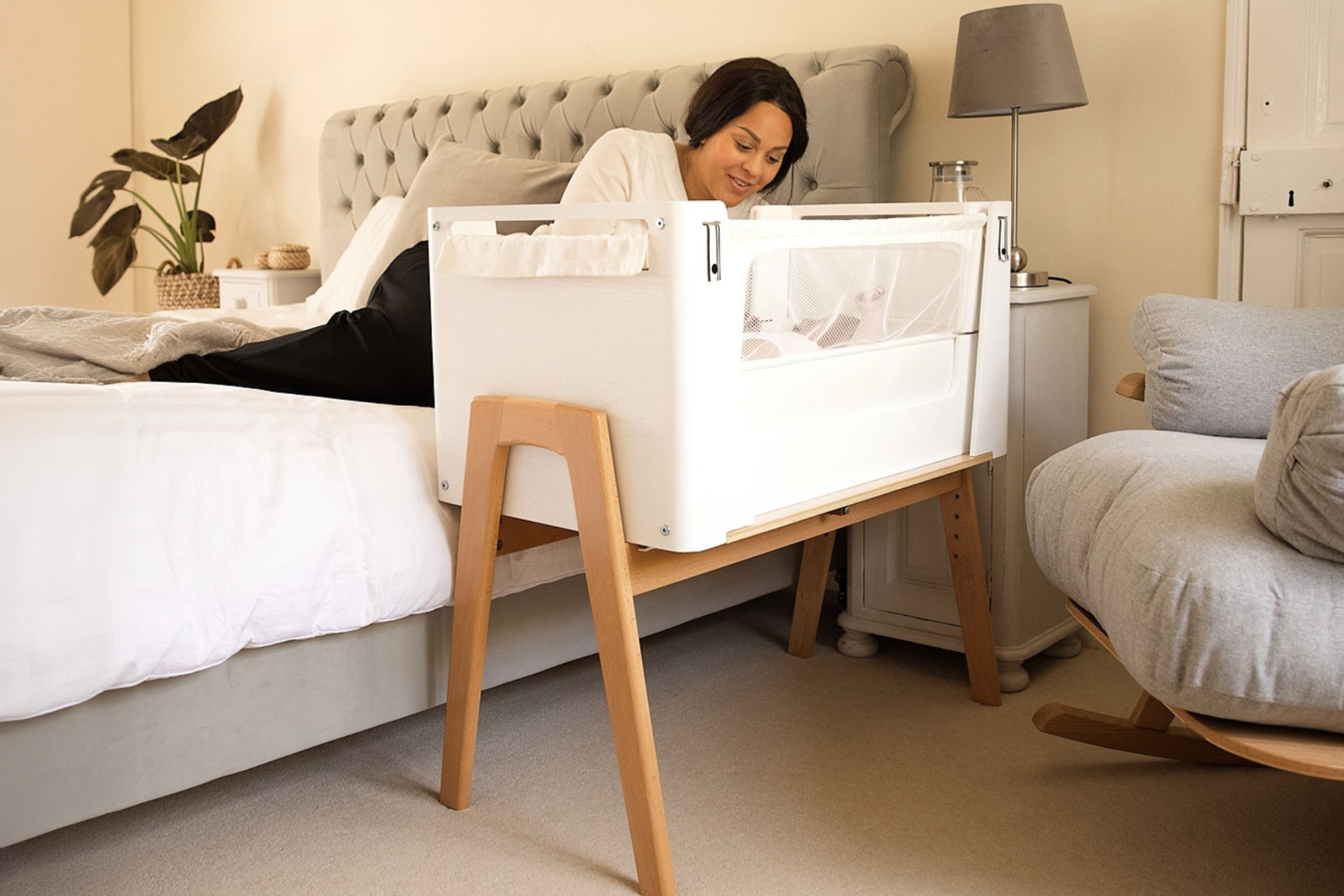 The Hera Bedside Co-Sleeping Crib next to parents bed while the mother reaches in to hold her little child.